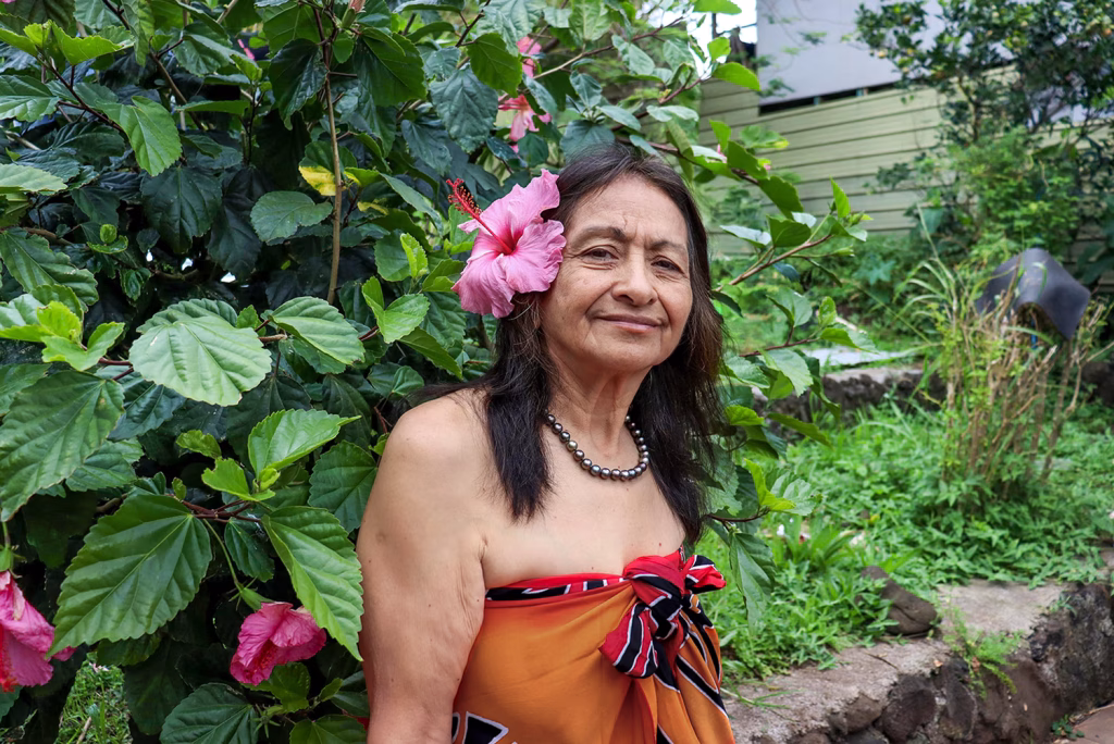 Orietta Rapu con una flor de hibisco en el cabello y un pareo naranja, frente a un fondo de follaje.